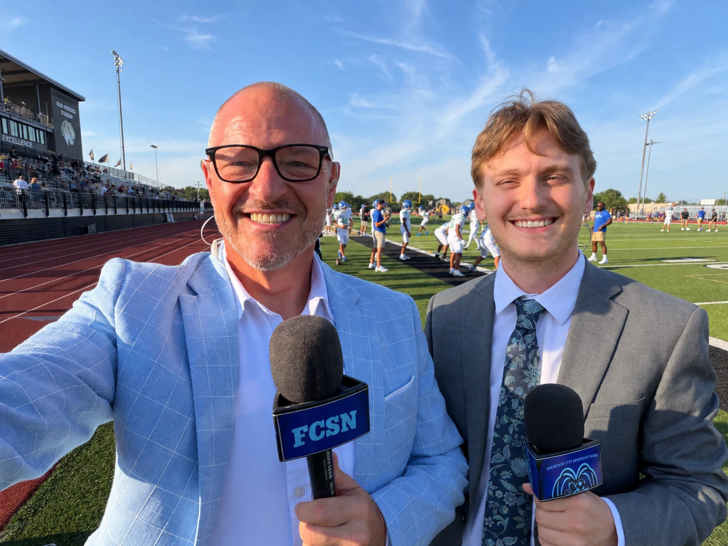 Chris Kamler and Brennen holding microphones beside a football field