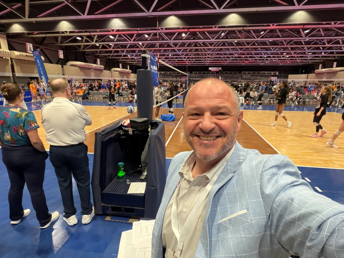 Chris Kamler smiling beside a volleyball court in a light blue jacket